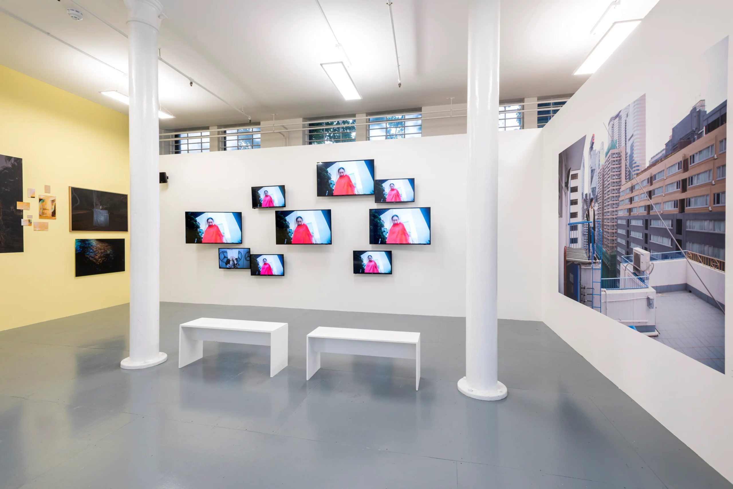 Image of a white-cube like gallery space, from a slight angle from the right. The far back wall is white and contains nine flat-screens of various sizes with an image of a dark-haired female figure, looking at the camera, dressed in bright red clothing. Screens are arranged in dynamic grid. There are two white, shinny cylinder pillars and two minimalistic white gallery benches in front of the wall. The wall to the left os painted in light yellow and has dark prints attached to it, while the wall to the right is white and is mainly covered with a large photograph of a person standing on a balcony in the middle of the image and a architectural daytime city-scape behind them. The floor of the space is grey and there are some light shinning from the windows behind the back wall.